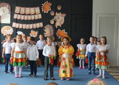 Dzieci w kolorowych strojach występują na scenie przed jesiennym tłem z dekoracjami.