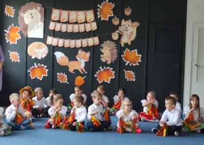 Grupa dzieci w białych koszulach siedzi na podłodze przed ozdobioną tablicą na Dzień Edukacji.