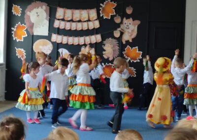 Dzieci na scenie z jesiennymi dekoracjami, występują w kolorowych strojach przedszkolnych.
