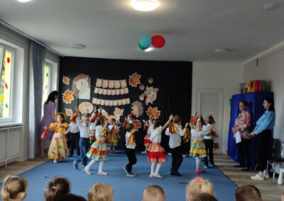 Dzieci tańczą na scenie w kolorowych strojach, jesienne dekoracje w tle, dorośli obserwują.