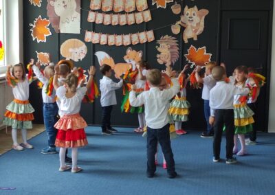 Dzieci tańczą w kolorowych strojach przy dekoracjach z jesiennymi motywami i zwierzętami.