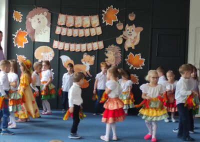 Dzieci w kolorowych strojach tańczą na scenie z jesiennymi dekoracjami, napis "Dzień Edukacji Narodowej".