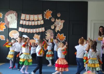 Dzieci w kolorowych strojach tańczą na scenie z jesiennymi dekoracjami.